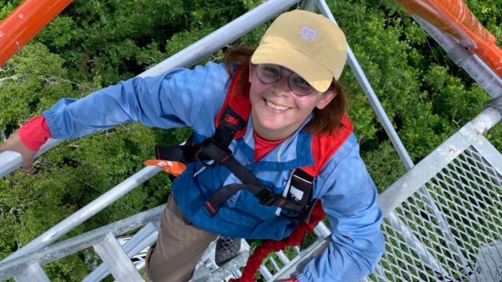 A woman wearing a baseball cap and glasses on a skywalk looking up at the camera