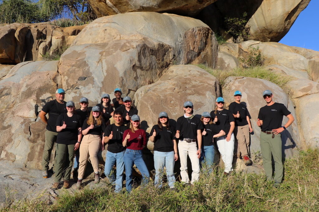 A group of students standing in front of really big rocks. Wearring the same black t-shirts and blue caps.